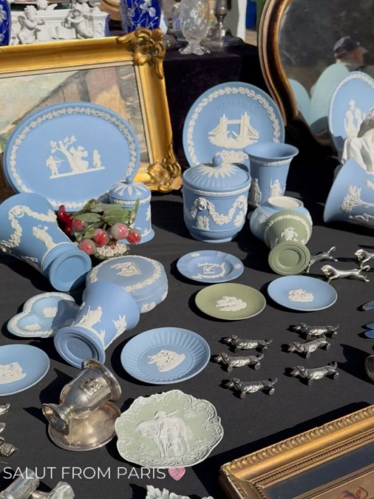 Blue and green Wedgwood style ceramics decorated with white classical figures displayed among silver miniatures and gilded frames at the Vanves Flea Market. Plates jars cups and small dishes crowd the table alongside a collection of tiny silver dog figurines.