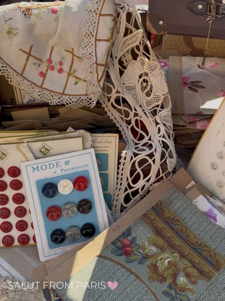 Vintage sewing and textile finds at a flea market stall including embroidered linens intricate white lace antique buttons and patterned fabric samples. Cards of colorful buttons include the text "MODE Parisienne" and a paper textile sample is marked "1923".