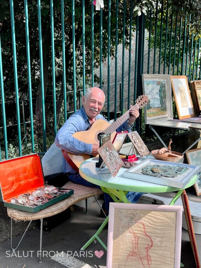 Man plays guitar and smiles in camera at Puces de Vanves
