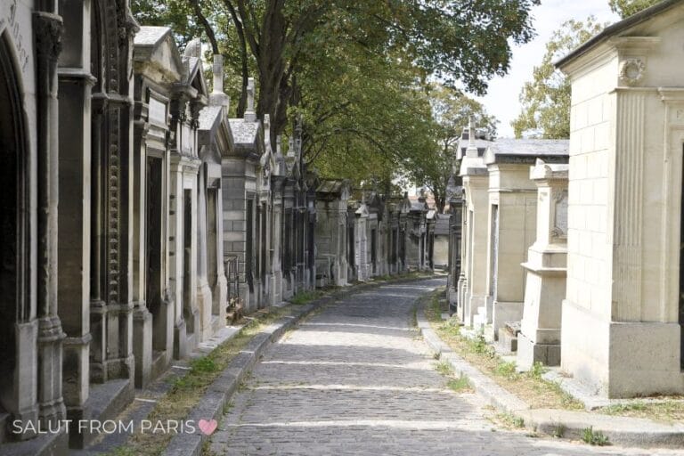 This image shows a peaceful cobblestone path flanked by historic mausoleums and tombstones in Pรจre Lachaise Cemetery, Paris. The sunlight filtering through the trees adds a serene touch to the timeless atmosphere of this renowned resting place.