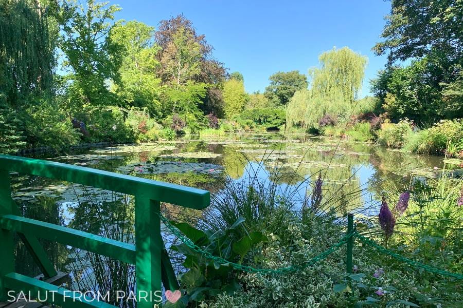 A serene view of the water lily pond in Giverny, France, with a green bridge in the foreground, surrounded by lush greenery, colorful flowers, and weeping willows under a bright blue sky.