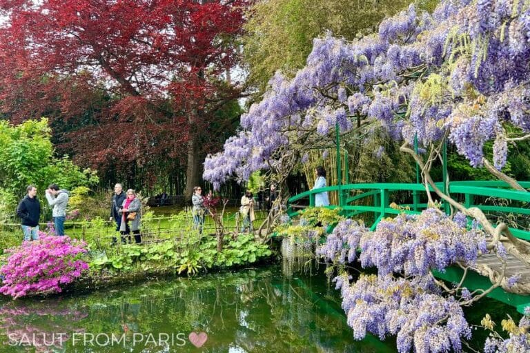 A serene garden scene in Giverny, France, featuring a green bridge draped in purple wisteria, a pond with lily pads, and vibrant trees in the background.