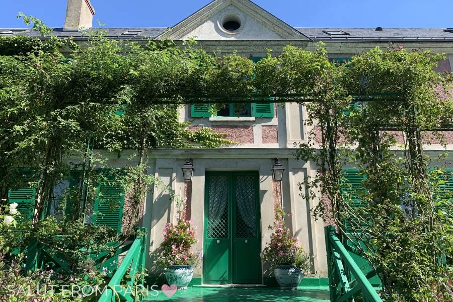 This image shows the front of Monet's House in Giverny, framed by green shutters and vines cascading down from an arched trellis. The bright green doors and pastel pink façade add to the picturesque charm of the famous artist's residence, surrounded by lush vegetation.