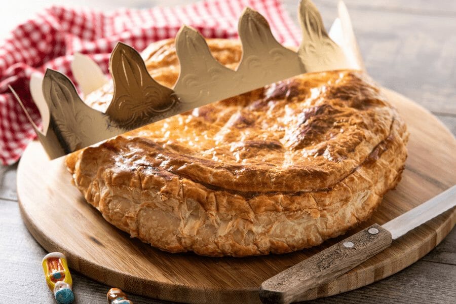 The image shows a traditional French Galette des Rois, or King Cake, resting on a wooden board. The cake, made of golden, flaky puff pastry, is topped with a decorative paper crown, symbolizing the Epiphany celebration. The Galette des Rois is typically filled with almond frangipane, and a small charm, or "fève," is hidden inside. The person who finds the fève in their slice is crowned king or queen for the day. A knife and a red checkered cloth are also visible, adding a rustic touch to the scene.