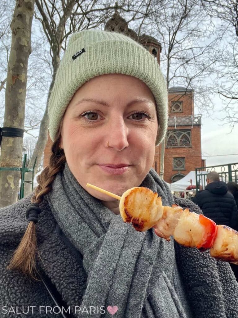 This image shows a woman bundled up in a beanie and scarf, holding a skewer of grilled scallops, likely at the Scallop Festival in Montmartre. Behind her, bare trees and a rustic brick building can be seen, creating a cozy, wintry outdoor setting. The text "Salut from Paris" with a heart is visible in the lower-left corner.