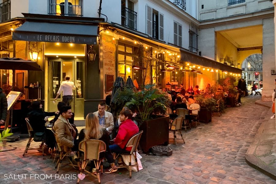 This image depicts a cozy outdoor dining scene at a Parisian café named "Le Relais Odeon." People are seated at small tables along a cobblestone street, enjoying drinks and conversation under warm string lights. The café’s façade features large windows and a dark awning, creating an inviting atmosphere. The street is narrow, framed by classic Parisian architecture, with a soft evening glow adding to the ambiance of the scene.