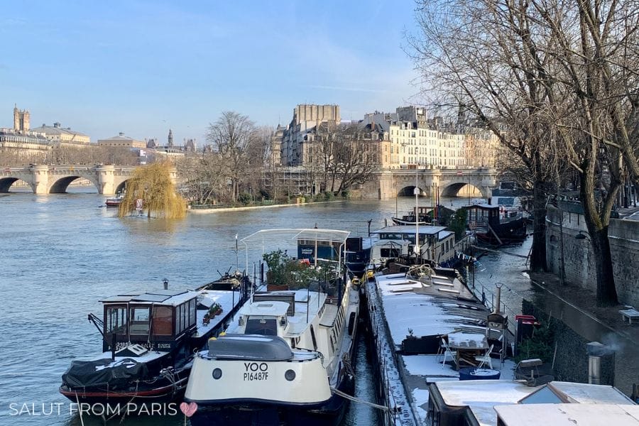 This image shows a serene winter scene along the Seine River in Paris, with houseboats lined up along the riverbank. Some of the boats have a light dusting of snow on their roofs. In the background, you can see a stone bridge crossing the river, with historic Parisian buildings and trees on the other side. The sky is clear and blue, and the bare trees hint at the cold season. The reflections in the water and the peaceful atmosphere capture the charm of Paris during winter.