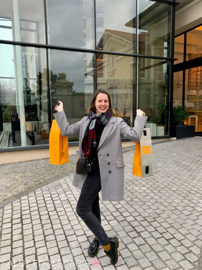 A woman, identified as Lena, poses excitedly outside the Veuve Clicquot Champagne House in Reims, holding two bright orange shopping bags from the renowned brand. She is dressed in a stylish gray coat, black jeans, and black loafers, accessorized with a plaid scarf and a small black crossbody bag. The modern glass-fronted building and cobblestone pavement add to the elegant setting, capturing the essence of a celebratory champagne experience.