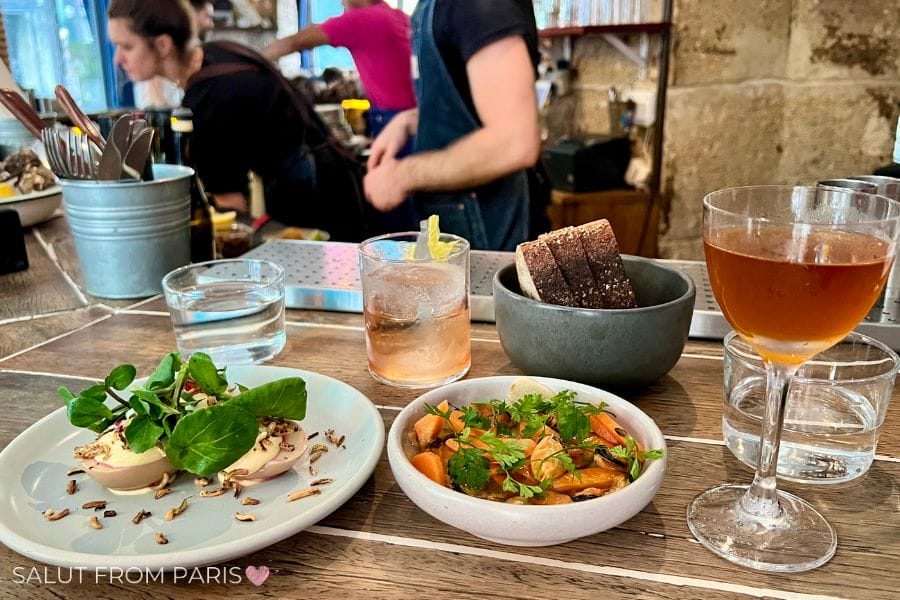 This image captures a beautifully presented meal at a Parisian restaurant. The table features several small dishes, including a plate of deviled eggs garnished with fresh greens, a bowl of hearty vegetables, and a bowl of rustic bread. Two drinks accompany the meal: a glass of water and a cocktail or glass of wine. In the background, you can see staff preparing food behind the counter, adding to the casual, lively atmosphere of the dining experience. 