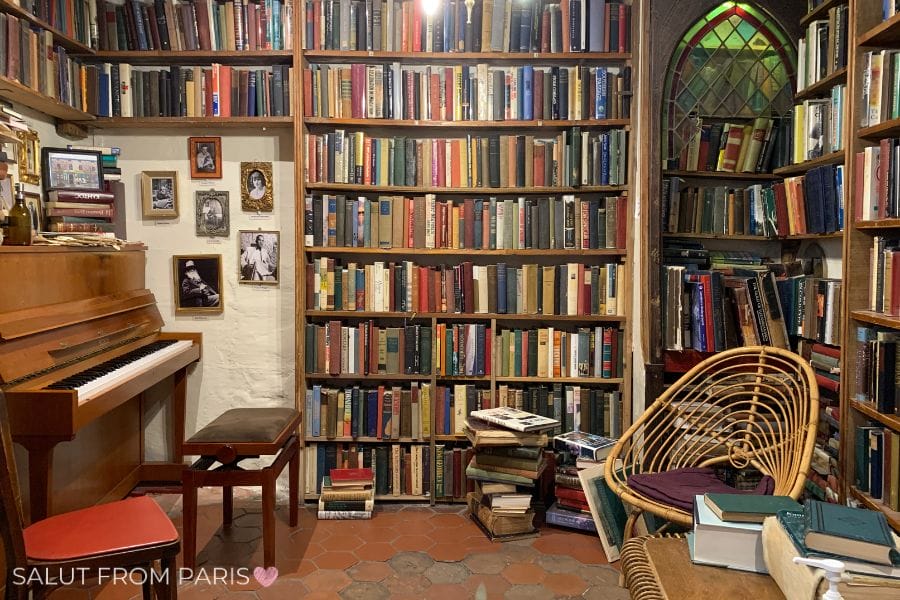 The image shows a cozy corner inside a bookstore, filled with shelves of colorful, well-worn books. A small wooden piano sits to the left, accompanied by a cushioned bench, while a wicker chair with a soft pillow is placed to the right. There are also a few stacks of books on the floor, adding to the literary charm of the space. The walls are adorned with black-and-white photos and framed art, contributing to the vintage, inviting atmosphere. The text "Salut from Paris" is seen at the bottom left with a small heart.