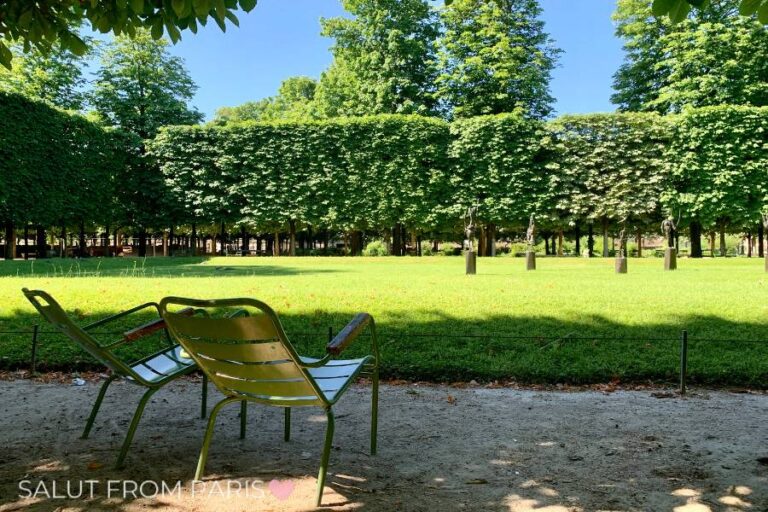 Two green metal chairs sit in the shade at the Jardin des Tuileries in Paris, facing a sunlit lawn bordered by perfectly manicured trees. In the distance, a row of classical statues stands among the greenery, adding to the elegance of the historic garden. The bright blue sky and lush foliage create a peaceful atmosphere.