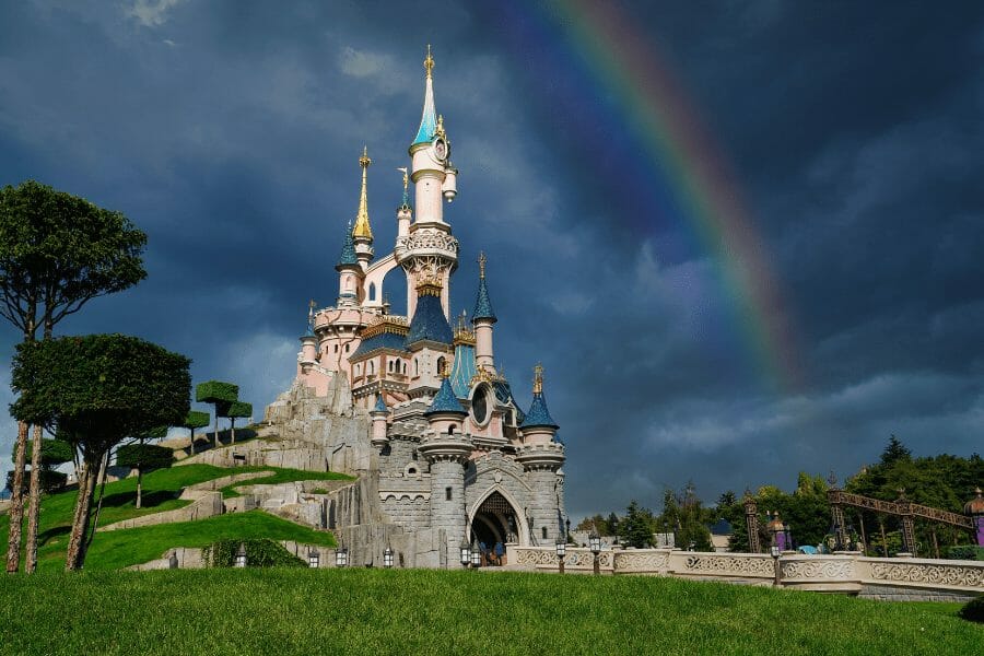 Sleeping Beauty Castle in Disneyland Paris under dramatic clouds and a glowing rainbow. The green lawn and whimsical towers create a fairytale setting. Just a short train ride from central Paris, the park is a favorite for families and Disney fans alike.