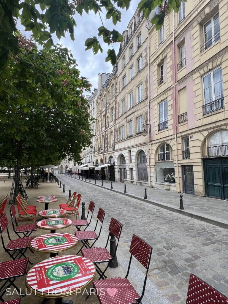 Outdoor café terrace with red bistro chairs and round tables on a quiet cobblestone street in Paris, leafy trees in spring and classic Haussmann buildings lining the road.