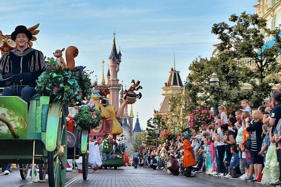 A colorful parade at Disneyland Paris features vibrant floats and performers dressed as beloved Disney characters, entertaining a large crowd. In the background, the iconic Sleeping Beauty Castle adds to the magical atmosphere. Spectators line the street, many capturing the moment on their phones, while the cheerful energy of the parade fills the scene. Lush trees and flowers along the street enhance the enchanting environment, creating a joyful and lively celebration of all things Disney.