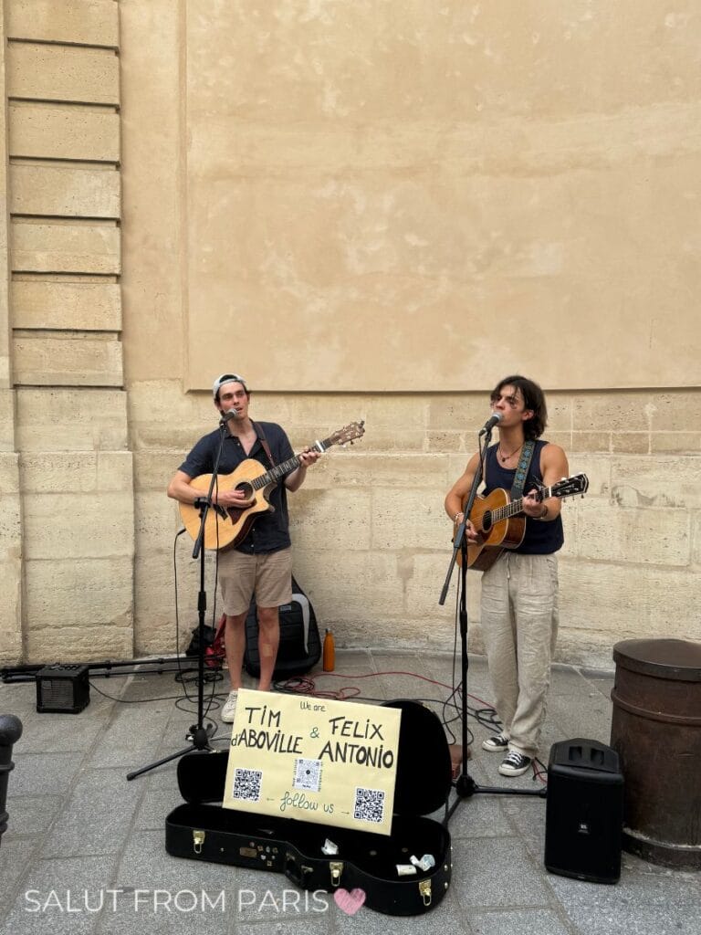 Two street musicians playing guitars and singing against a stone wall with a sign reading "We are Tim d’Aboville & Felix Antonio follow us" and text "SALUT FROM PARIS" capturing live music in June in Paris.