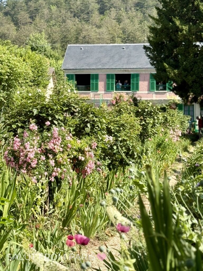 Lush flower garden in full bloom with a pastel pink house and green shutters behind it showing the beauty of gardens to visit during June in Paris with text "SALUT FROM PARIS".