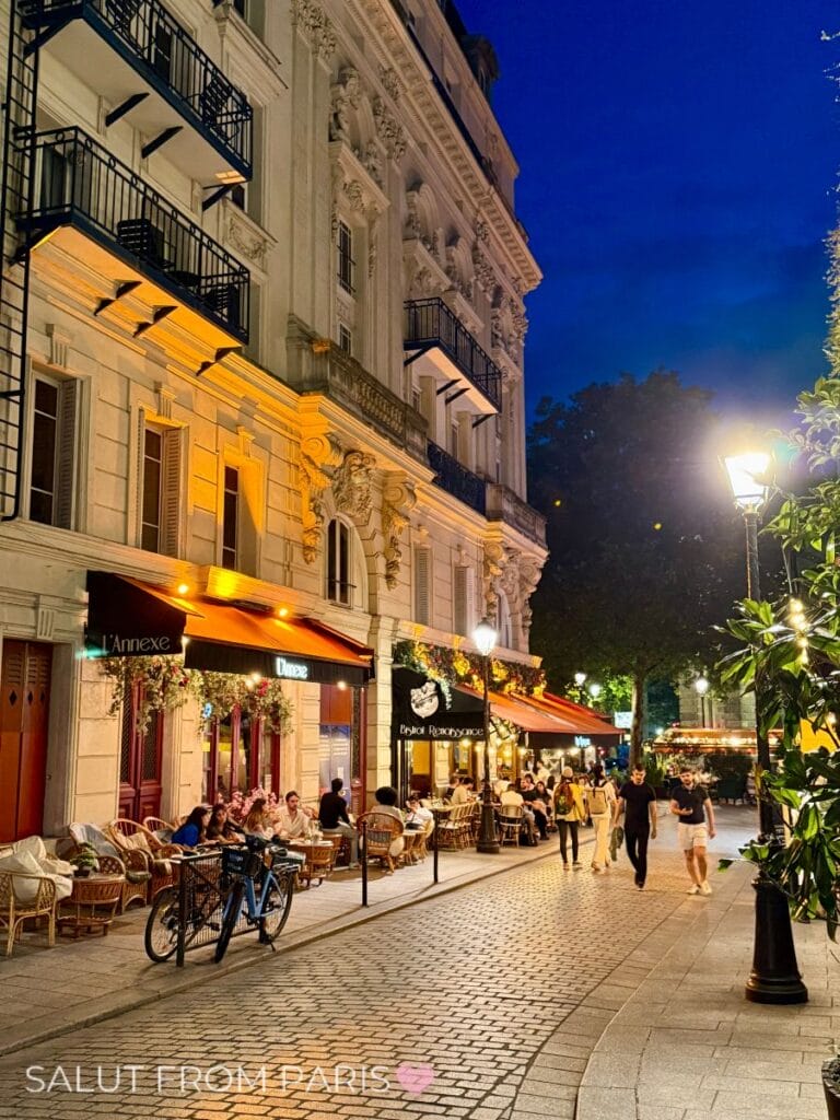 Paris by night with cobble stones lanes and warm street lanterns illuminating the scenes. People sitting in cafés and the Haussmann buildings to the left are wamly illuminated