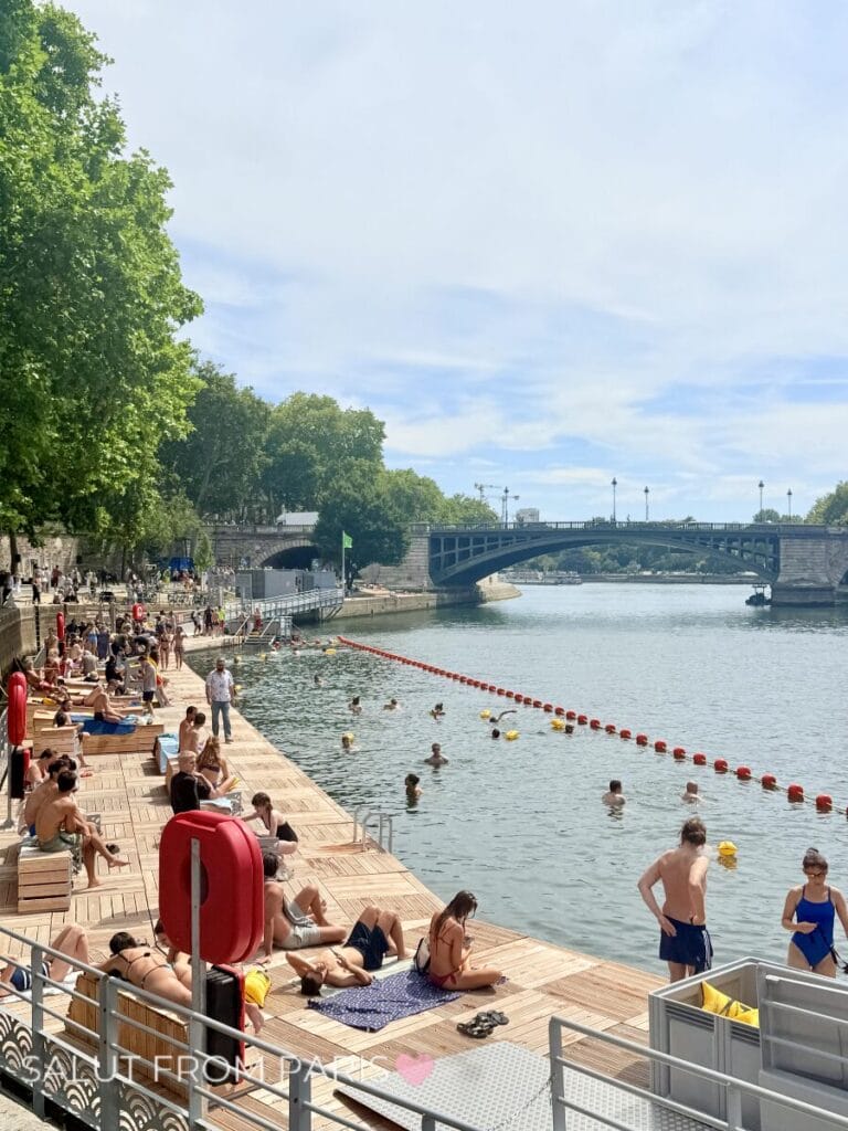 Swimmers cool off in a designated river swimming area along the Seine while others sunbathe on a wooden deck beside the water. The lively waterfront scene highlights how locals and visitors enjoy warm weather in July in Paris.