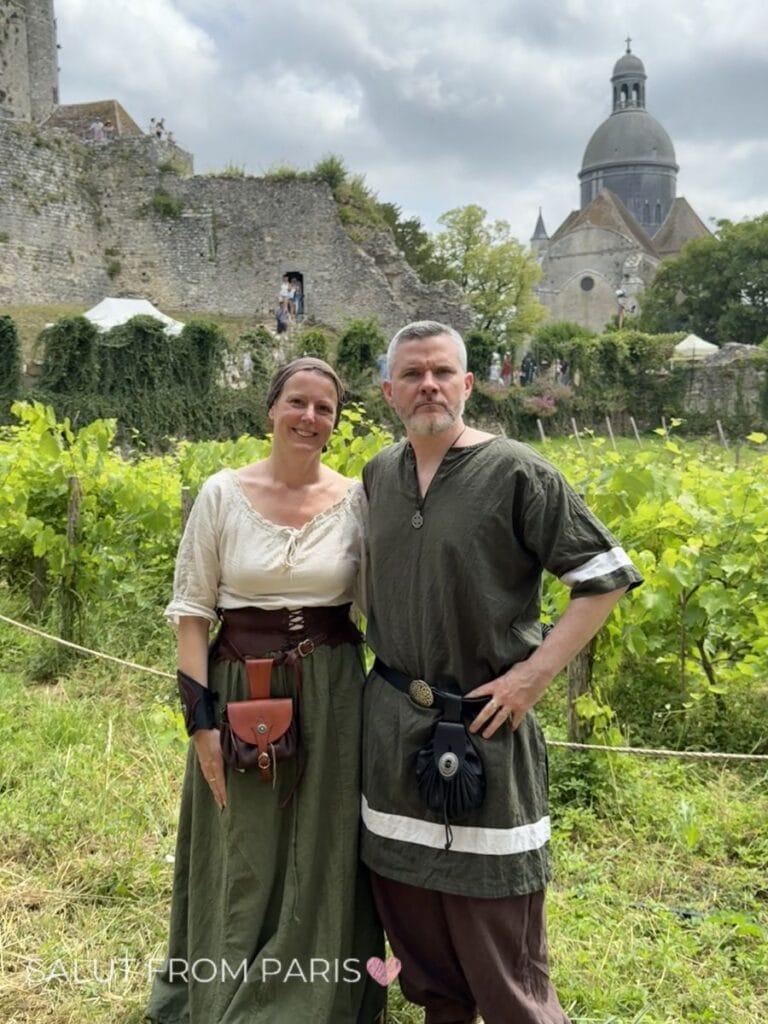Couple dressed in medieval style clothing standing in a vineyard with historic stone ruins and a domed building behind them showing a unique cultural experience near Paris in June with text "SALUT FROM PARIS". If you are wondering what to do in Paris in June, Provins is a good idea for a side trip