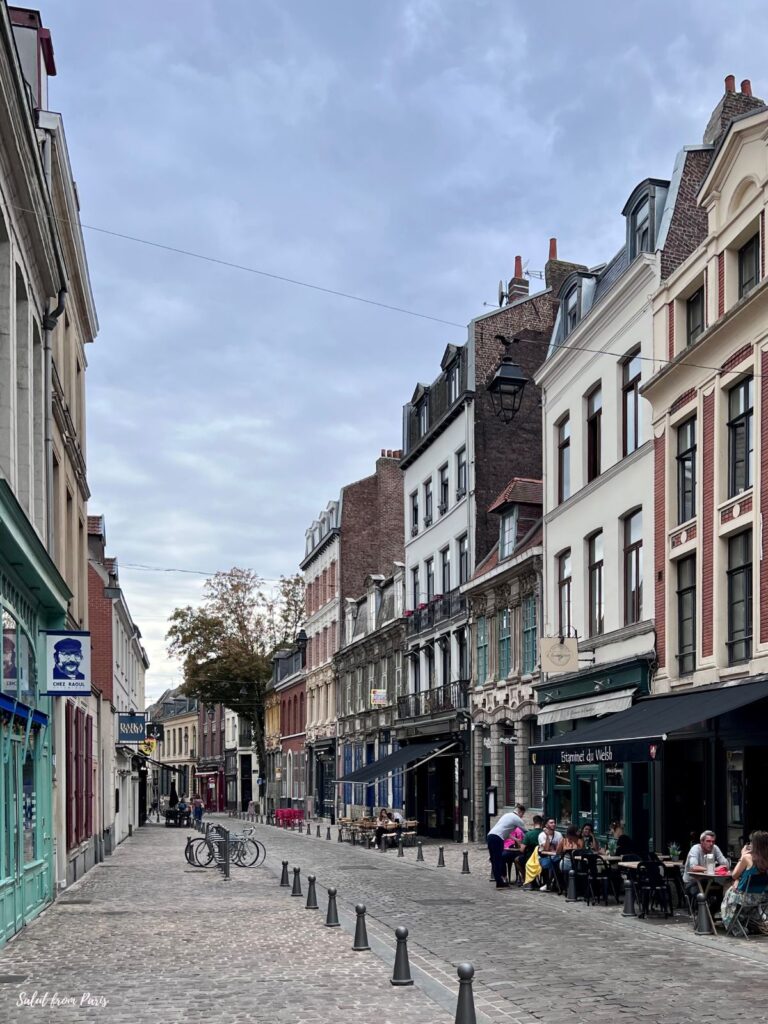 A quiet cobblestone street in Lille lined with historic brick buildings and small cafés. People sit outside enjoying drinks under soft morning light. The charming mix of architecture and relaxed atmosphere captures the essence of northern France.
