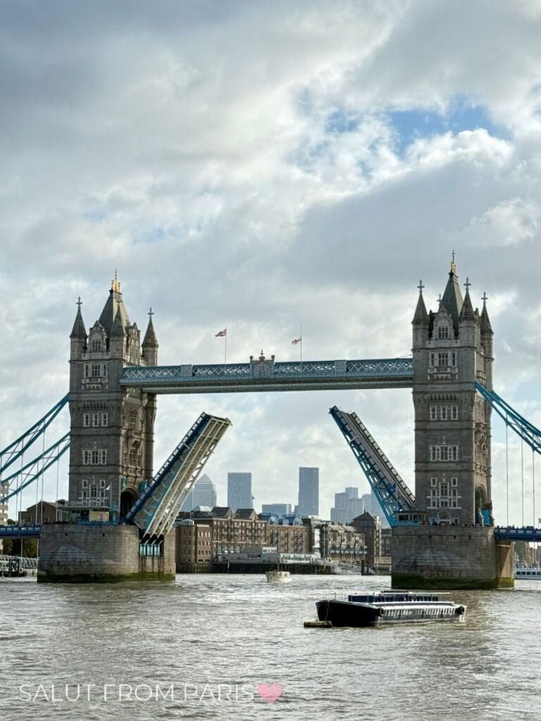 The famous Tower Bridge in London stands over the River Thames with its bascules raised to let a boat pass. The cloudy sky and distant city skyline create a dramatic backdrop. This iconic landmark is a highlight for anyone visiting London on a day trip from Paris.
