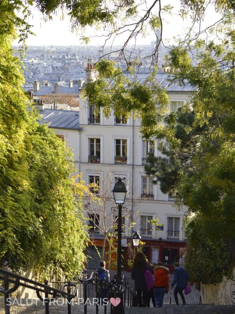Stone steps descend through a leafy Montmartre hillside toward classic Paris apartment buildings and rooftops below. A vintage streetlamp stands in the center while visitors make their way down the staircase. This quiet neighborhood scene is a charming glimpse of Paris in Fall.