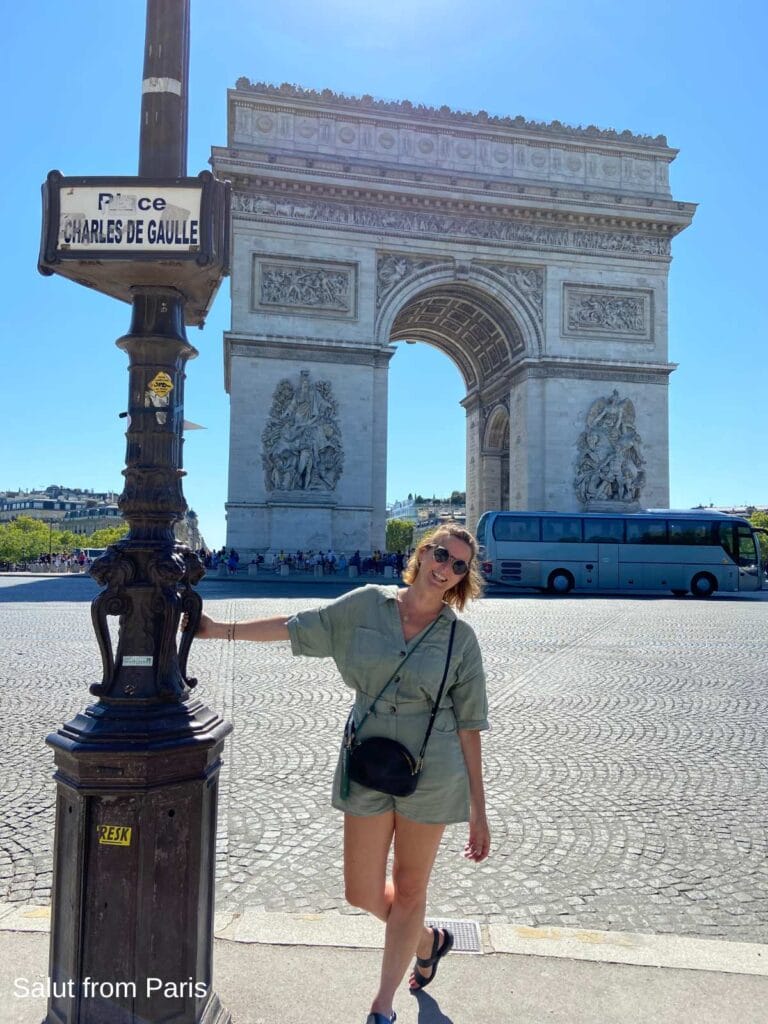 Woman in a light green romper standing beside a Paris street sign at Place Charles de Gaulle, with the Arc de Triomphe rising in the background on a bright August day.