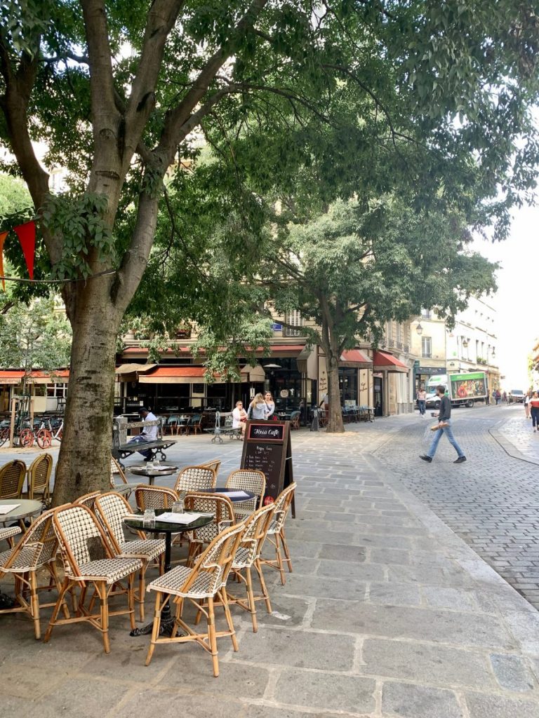 Wicker cafรฉ chairs and small tables sit beneath large shade trees along a quiet cobblestone street lined with neighborhood restaurants. A few pedestrians walk through the relaxed square surrounded by classic Paris architecture. Outdoor cafรฉs like this are perfect for enjoying Paris in September.