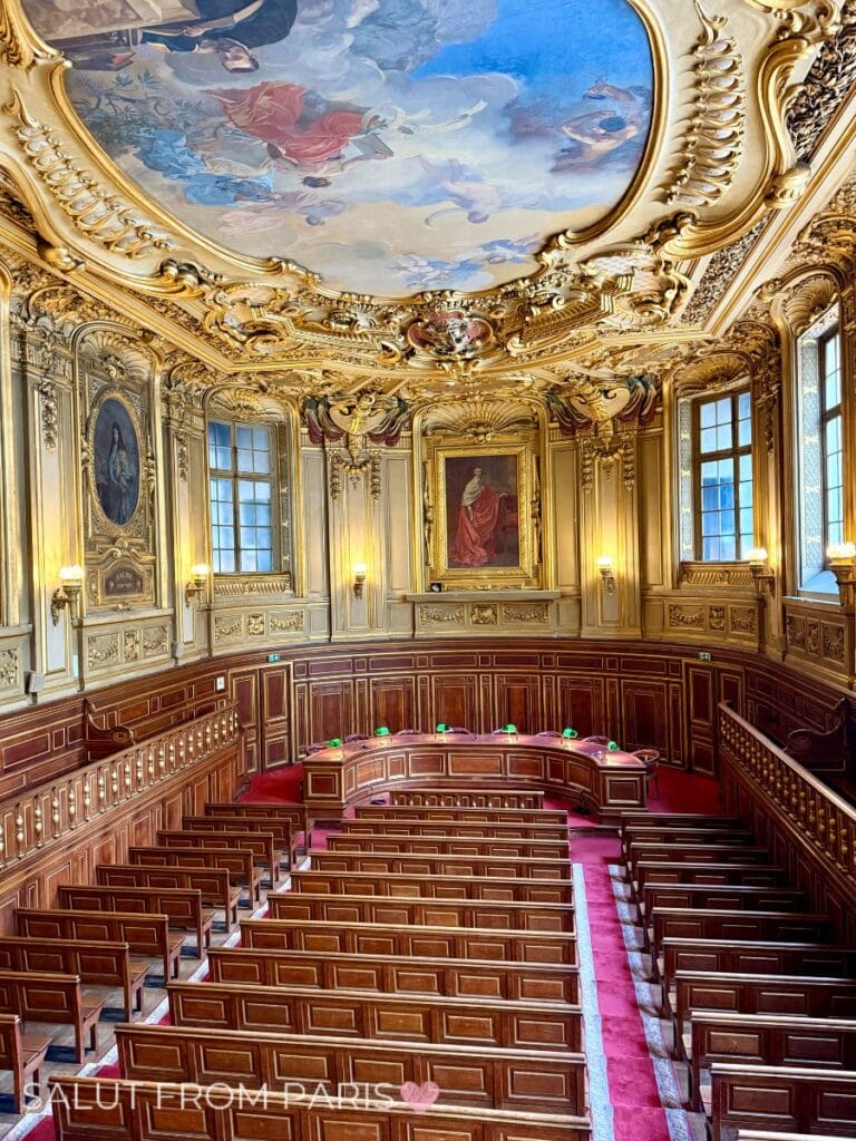 Ornate historic courtroom with polished wooden benches, carved paneling, gilded walls, and an elaborate painted ceiling. Tall windows fill the grand chamber with natural light while decorative artwork surrounds the elevated judges bench. The richly preserved interior highlights the opulent architecture found in Paris.