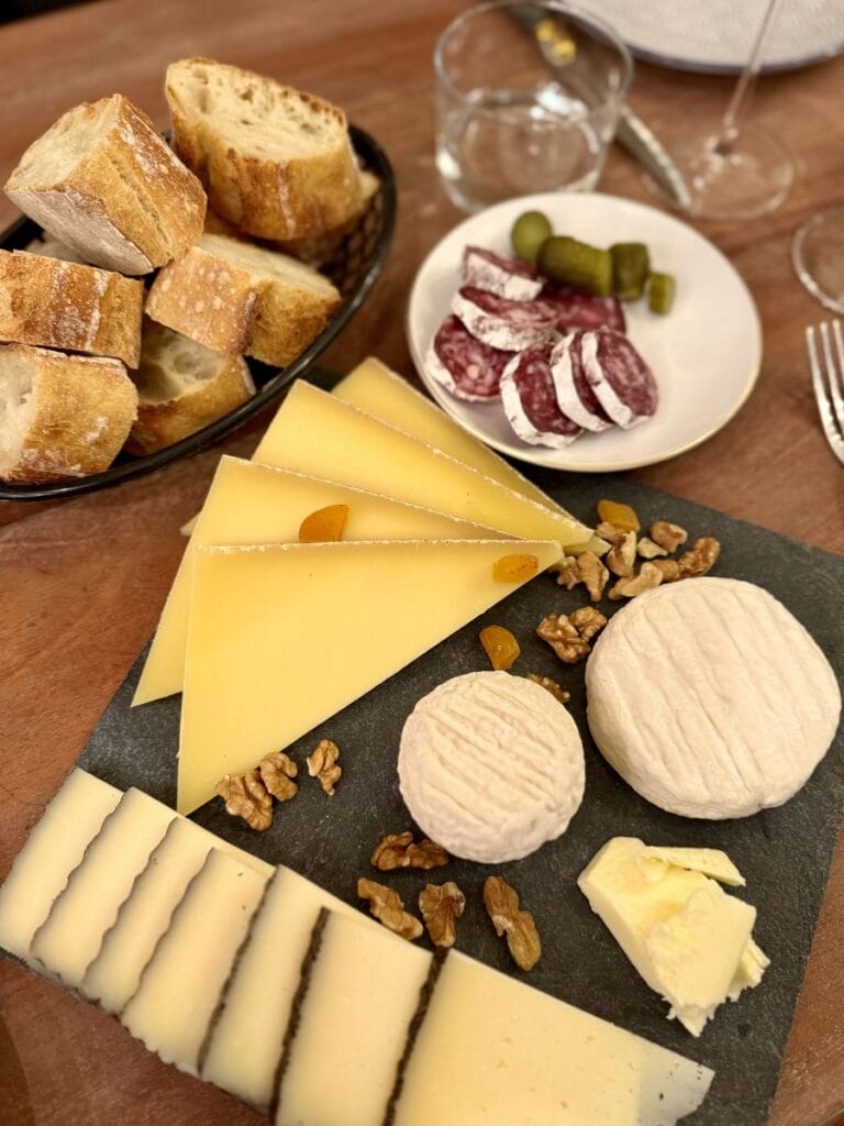 cheese platter with different kind of cheeses and a bowl with fresh baguette. There's also a little bowl with cold cuts and cornichons. everything is placed on a wooden table