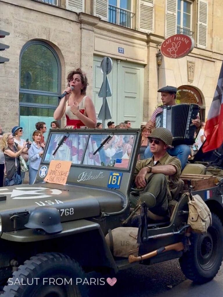 A singer performs from a vintage military jeep while an accordion player entertains the crowd during a lively street event. Spectators gather along the sidewalk outside historic Paris buildings, creating a festive atmosphere. This local celebration showcases the vibrant culture of Paris in September.