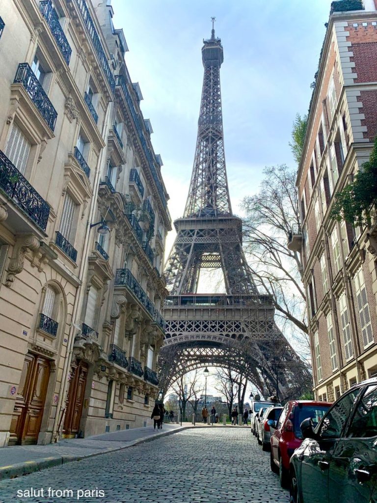 The image shows a stunning view of the Eiffel Tower framed by classic Parisian buildings on a narrow cobblestone street. The buildings on either side feature ornate balconies and elegant stone facades, typical of the architecture in this historic part of Paris. The Eiffel Tower rises majestically at the end of the street, its iron latticework contrasting with the stone structures. The scene captures the timeless beauty of Paris, blending iconic landmarks with the charm of everyday city life.