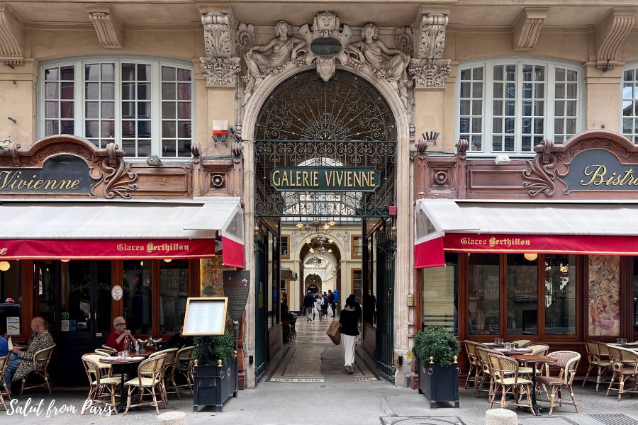 The entrance to Galerie Vivienne in Paris, framed by ornate stonework and sculptures above a large, decorative wrought-iron archway. On either side, red-awning cafes with outdoor seating add to the Parisian charm, while people walk through the elegant passageway inside.