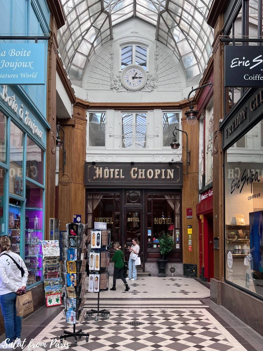 The entrance to Hรดtel Chopin inside a covered passageway in Paris, marked by vintage signage and a decorative clock above with a glass-domed ceiling. A display rack of postcards stands in front of nearby stores, while a few visitors browse and walk towards the hotelโs inviting wooden doors.