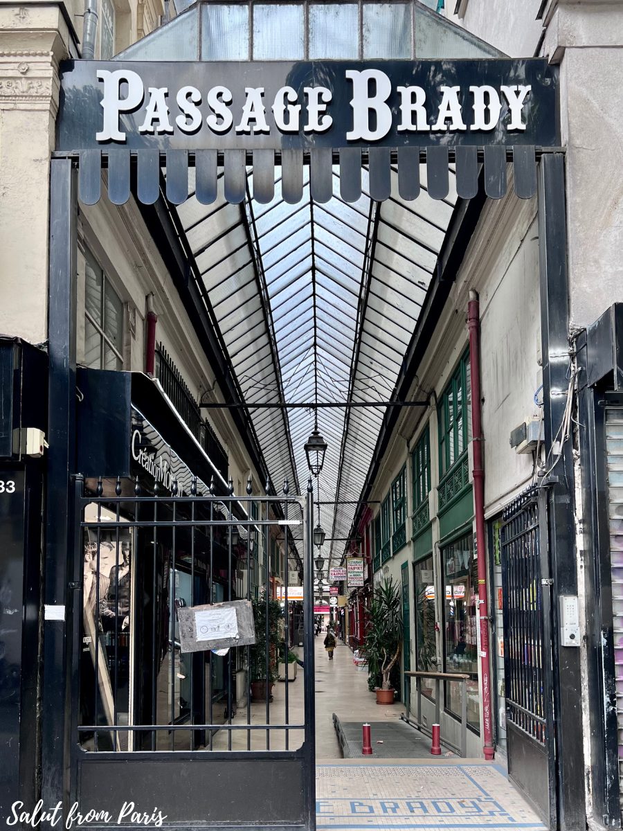 The entrance to Passage Brady, a covered arcade in Paris, featuring a black sign with white lettering and a metal gate. Inside, the passage is lined with shops and decorated with plants, illuminated by hanging lanterns under a glass roof. The tiled floor at the entrance reads โPassage Brady,โ welcoming visitors into the bustling walkway.