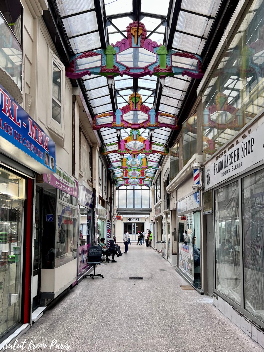 A colorful Parisian arcade with vibrant geometric designs hanging from the glass ceiling, adding a lively touch to the space. The walkway is lined with small shops, including a barber shop and a salon, with customers casually seated outside. In the distance, the sign for a hotel is visible, giving the passage a mix of local business activity and a welcoming, everyday atmosphere