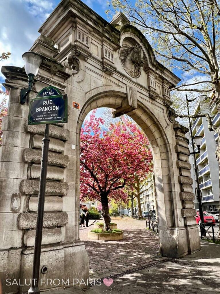 This image shows a charming stone archway on Rue Brancion in Paris, framing a blooming cherry blossom tree in the background. The arch, with its intricate architectural details, provides a glimpse into a quaint square where the vibrant pink blossoms of the tree contrast beautifully against the gray stone and blue sky. A street sign for "Rue Brancion" is visible on the left, adding a sense of location to the picturesque scene