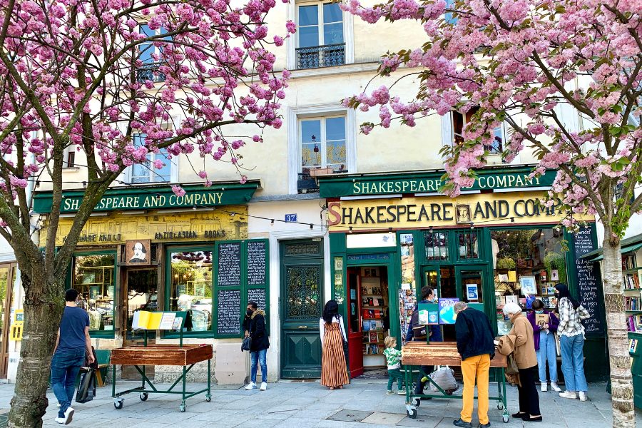This image shows the famous "Shakespeare and Company" bookstore in Paris, framed by two blooming cherry blossom trees in the foreground. The store's green exterior and vintage signage are clearly visible, with people browsing books displayed on tables outside. The pink blossoms above create a picturesque, inviting scene, and the warm, springtime atmosphere adds to the charm of this iconic literary landmark.