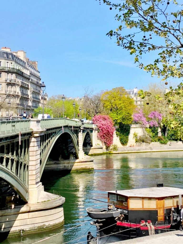 This image depicts a peaceful riverside scene in Paris, with a green iron bridge stretching across the calm waters of the Seine. A houseboat is moored in the foreground, while vibrant pink and green foliage lines the riverbanks, adding a pop of color to the otherwise historic cityscape. The background features classic Parisian buildings, their balconies and windows overlooking the water. The bright blue sky and serene river create a picturesque and tranquil setting, characteristic of a spring day in the city.