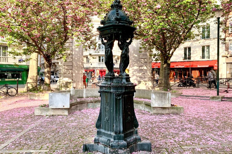 This image depicts a quaint public square with a dark green, ornate fountain at its center, surrounded by pink cherry blossom trees in bloom. The ground is carpeted with fallen pink petals, giving the scene a soft, enchanting atmosphere. In the background, you can see shops with bright red awnings and a few pedestrians walking by, while the surrounding trees and blossoms create a calm, picturesque urban setting. The fountain itself is a classic Parisian drinking fountain with statues supporting its dome.