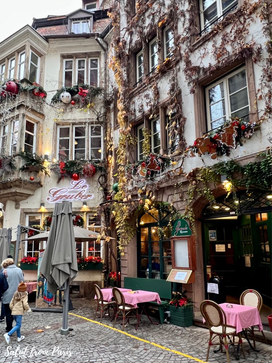 The image shows a charming alleyway in a historic European city, with buildings adorned in festive decorations for the holiday season. The façades are covered in ivy, some of which has turned brown for the winter, while balconies and windows are embellished with garlands, red bows, and Christmas ornaments. The cozy restaurant "Le Gruber" features pink tablecloths on its outdoor tables, inviting diners to enjoy a meal in this picturesque setting. The cobblestone street and warm lighting enhance the quaint, festive atmosphere.