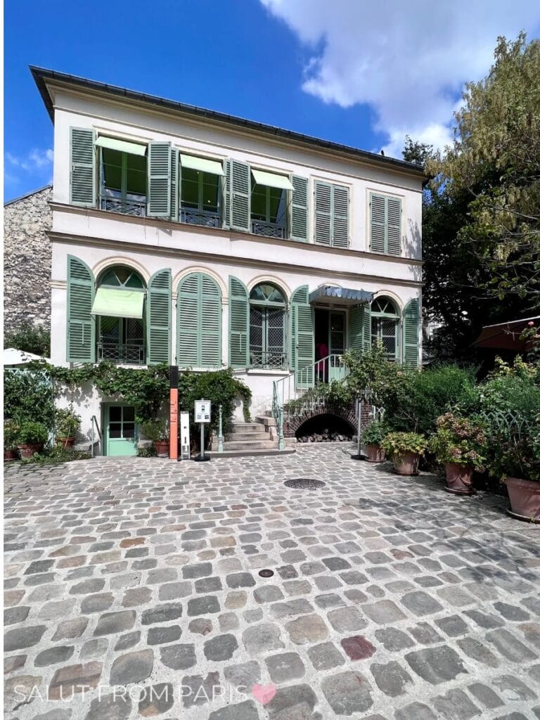 The image shows a charming two-story house with light green shutters and awnings, located on a cobblestone courtyard. The house has arched windows and a small set of stairs leading to the entrance, surrounded by potted plants and greenery. The bright blue sky and leafy trees in the background add to the peaceful, picturesque ambiance of this inviting space.