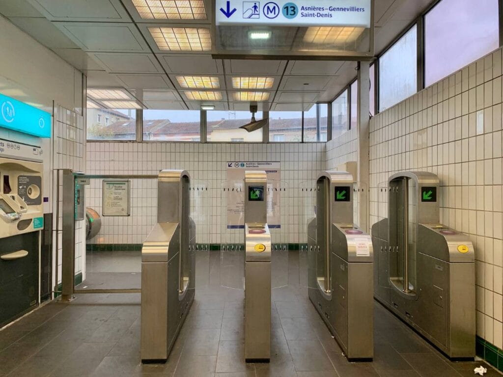 An entrance area of a Paris Metro station featuring several turnstiles with green arrow indicators for entry. On the left, there is a ticket machine under a blue 'Tickets' sign. The walls are tiled in white, and a sign above points towards Line 13, with directions to Asnières–Gennevilliers and Saint-Denis. The station is clean and well-lit with large windows providing natural light.