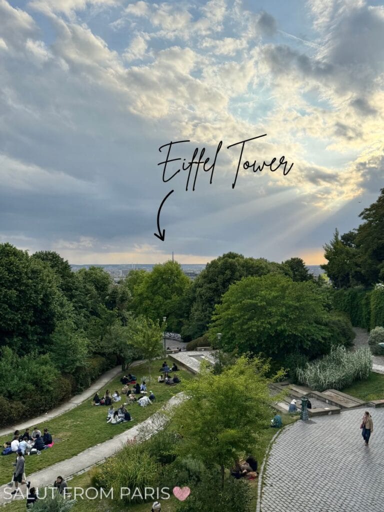 View from Parc de Belleville in Paris at sunset, with people sitting on grassy terraces and pathways. The Eiffel Tower is visible on the horizon, marked with handwritten text and an arrow pointing to it against a dramatic sky with rays of sunlight breaking through the clouds.
