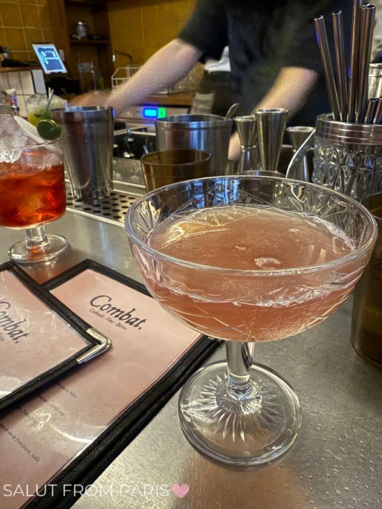 Close-up of a pink cocktail served in a cut-crystal coupe glass on the bar counter at Combat, a popular cocktail bar in Belleville, Paris. In the background, a bartender prepares drinks, and menus with the Combat logo are visible on the counter.