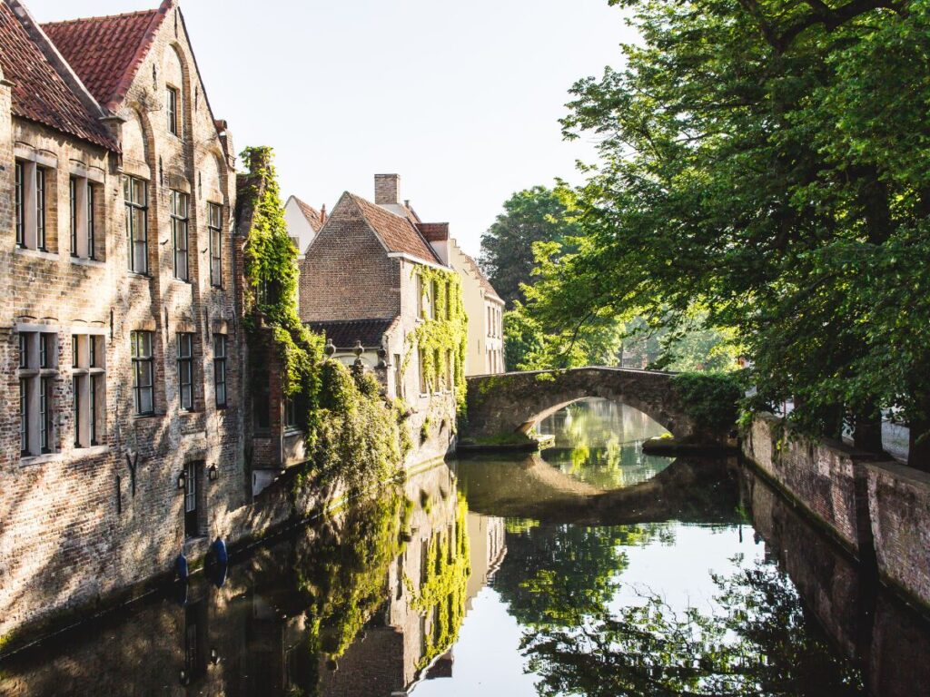 A peaceful canal in Bruges lined with ivy-covered brick houses and shaded by tall green trees. The small stone bridge reflects perfectly in the calm water, creating a postcard-perfect scene. Bruges’ medieval charm and tranquil waterways make it one of Belgium’s most beautiful towns.
