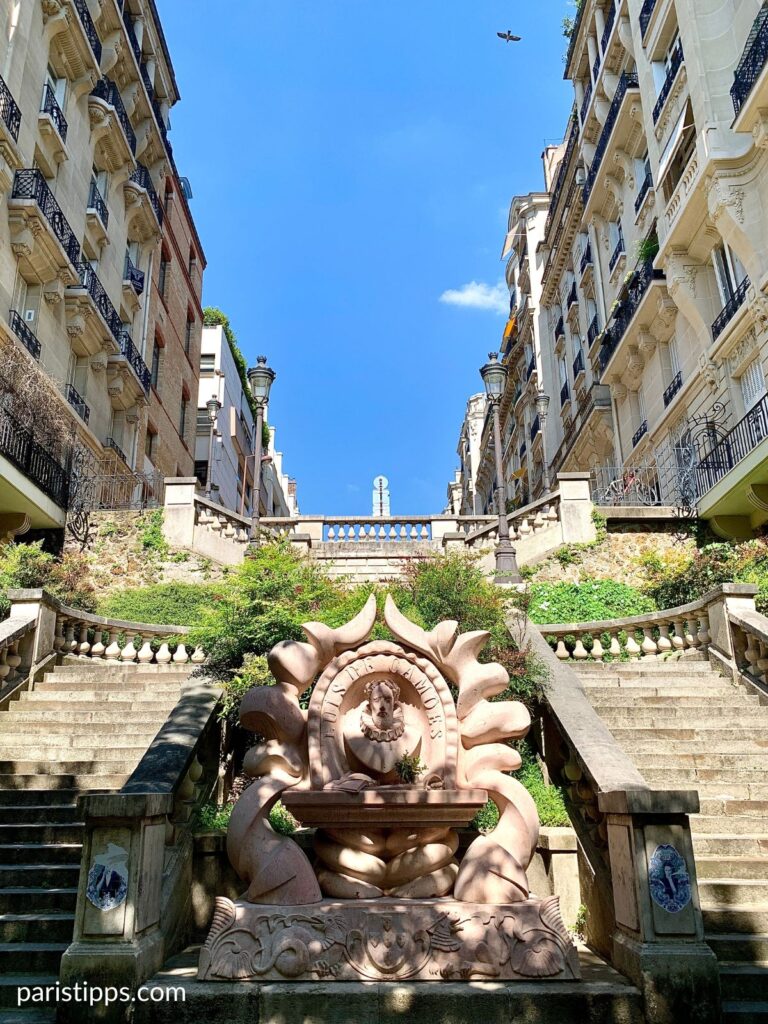 Ornate fountain and staircase in a Paris arrondissement surrounded by greenery and historic buildings climbing up the hill. Decorative stonework and lampposts lead the eye upward between the buildings.