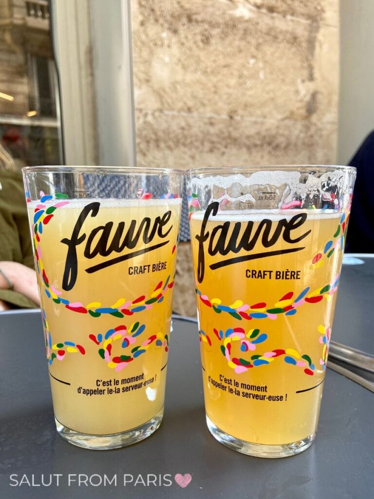 Two glasses of beer on a café table in a Paris arrondissement with colorful branding reading "fauve craft bière" and "C'est le moment d'appeler le.la serveur.euse !" Foam rests at the top as people sit nearby.