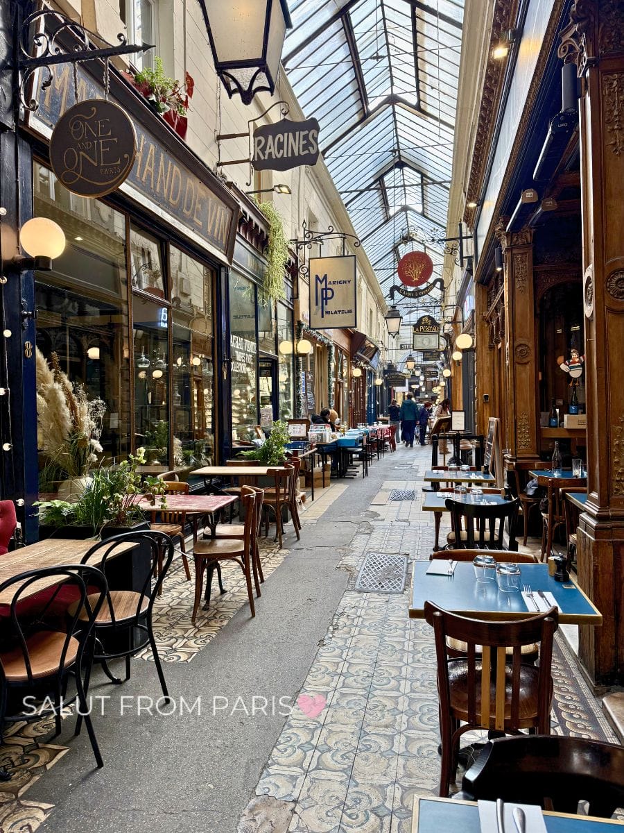 Covered shopping passage in a Paris arrondissement lined with cafés and boutique storefronts. Small tables and chairs sit along a tiled walkway under a glass ceiling with hanging signs including "One One Paris," "Racines," and "Marigny Philatelie," while a few people stroll through the corridor.