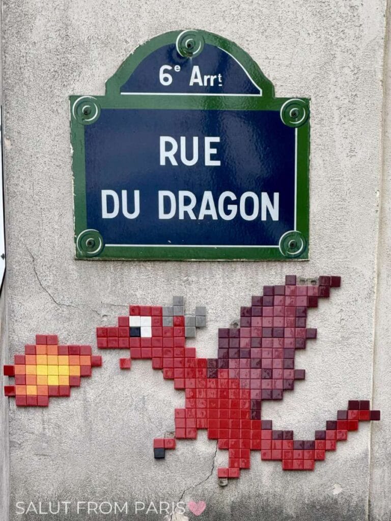 Blue Paris street sign reading "6e Arrt. Rue du Dragon" mounted on a stone wall in the 6th arrondissement, with a pixel-style red dragon mosaic breathing fire below it. The scene highlights a charming detail of arrondissements in Paris with playful street art beneath the official sign.
